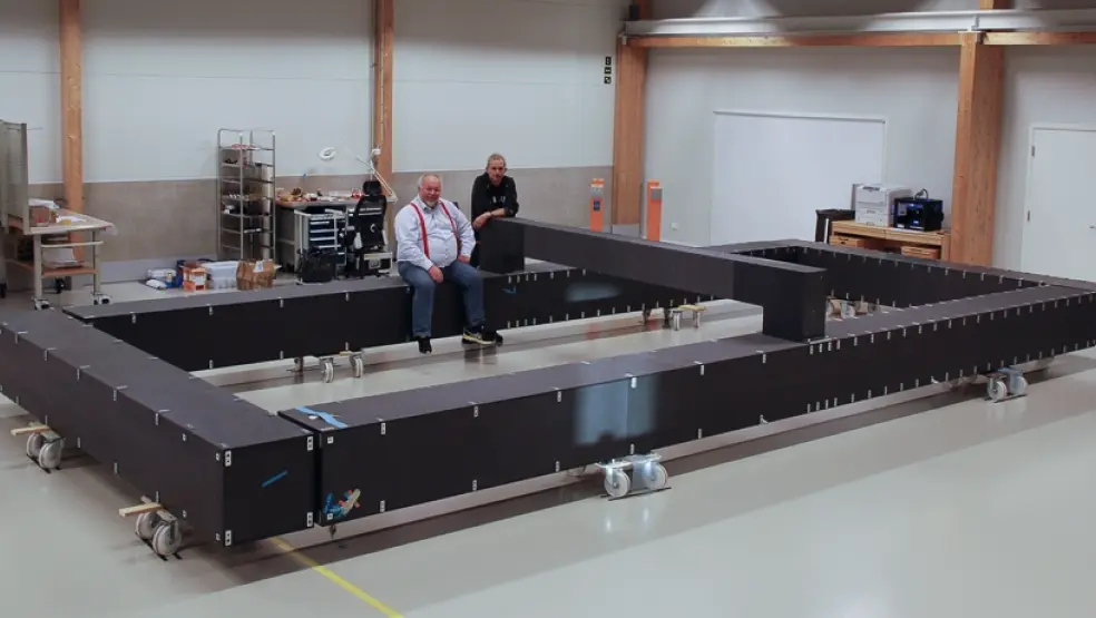 Two people sit on a large, rectangular metal frame in a workshop.