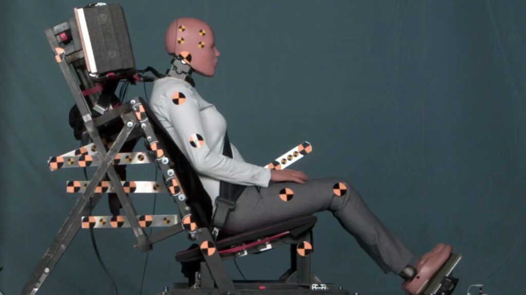 The image shows a crash test dummy seated in a vehicle, ready for a crash test. The dummy is equipped with various sensors and instruments to measure the impact and effects of the crash. The background appears to be a controlled testing environment, likely a laboratory or testing facility. The purpose of the image is to illustrate the safety testing procedures and the use of crash test dummies in evaluating vehicle safety.