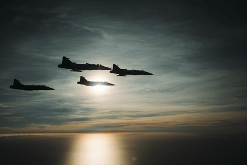 The image depicts a dramatic aerial scene at sunset. Several silhouetted fighter jets or aircraft are visible in the sky, flying in a formation against a backdrop of vibrant orange and pink hues.